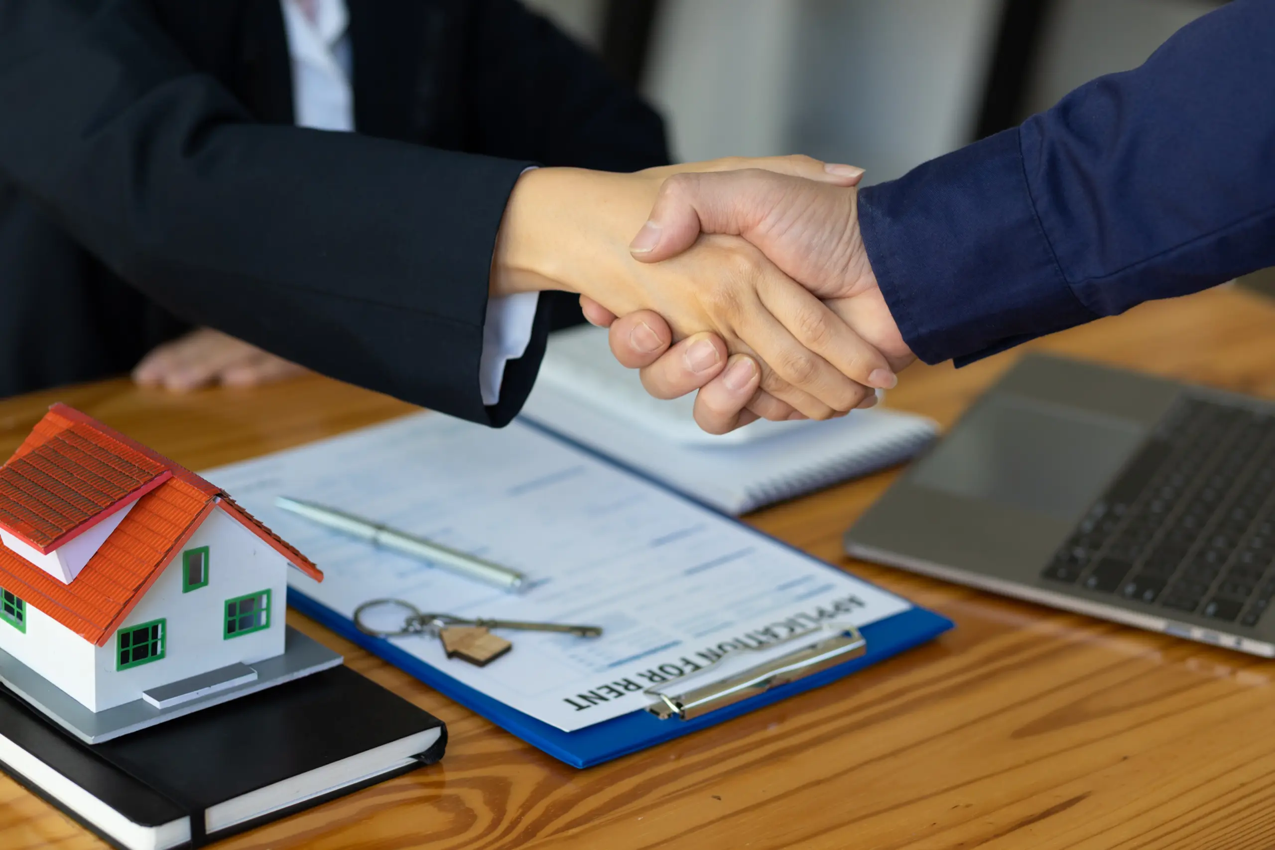 Zwei Personen geben sich die Hand über einem Tisch mit Immobilienvertrag, Hausschlüssel und Hausmodell. - Deutsch Marketing